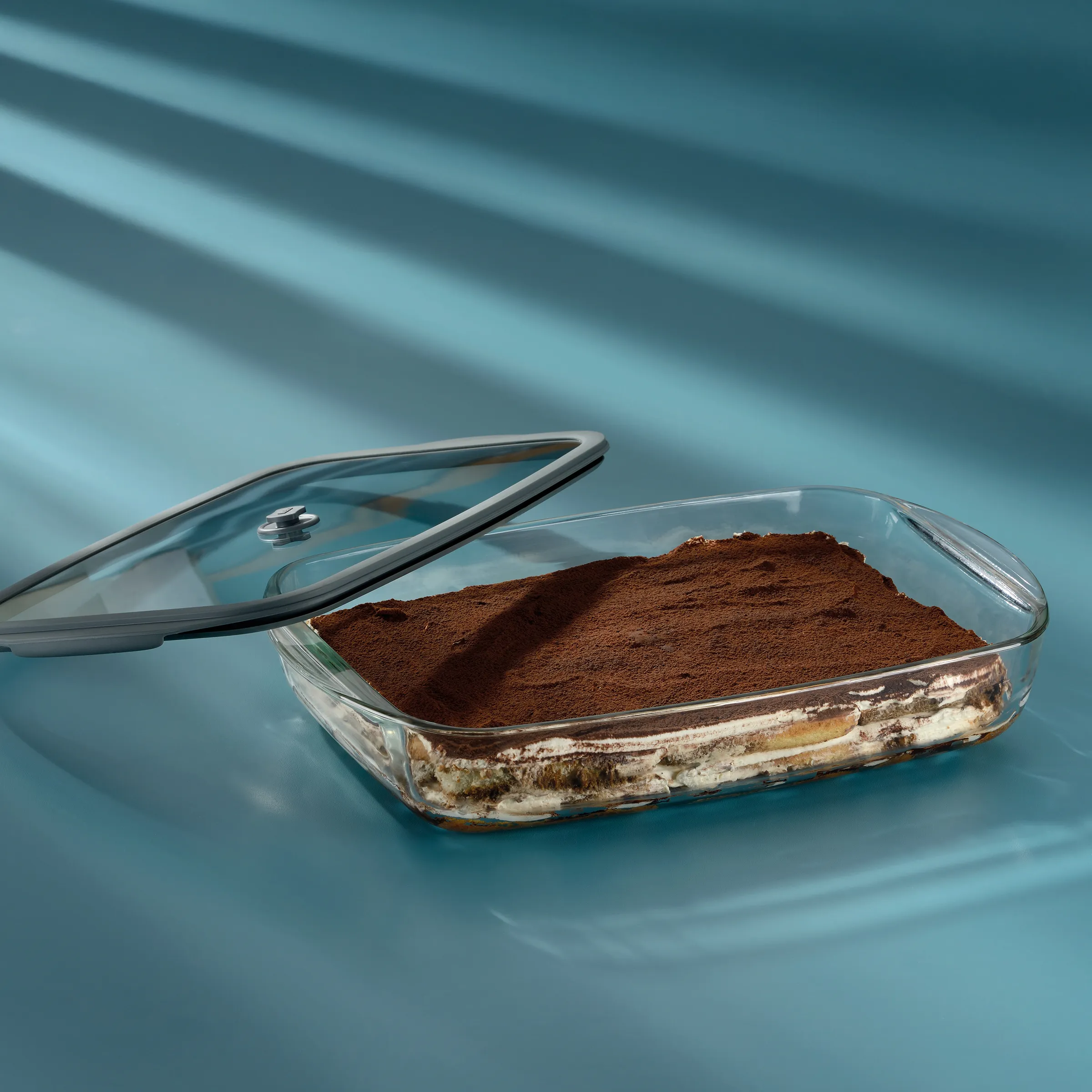 Glasauflaufform mit Tiramisu-Schichten und Kakaopulver auf blauem Hintergrund mit Glasdeckel