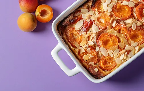 Aprikosen-Mandelkuchen in weißer Auflaufform mit frischen Aprikosen auf violettem Hintergrund