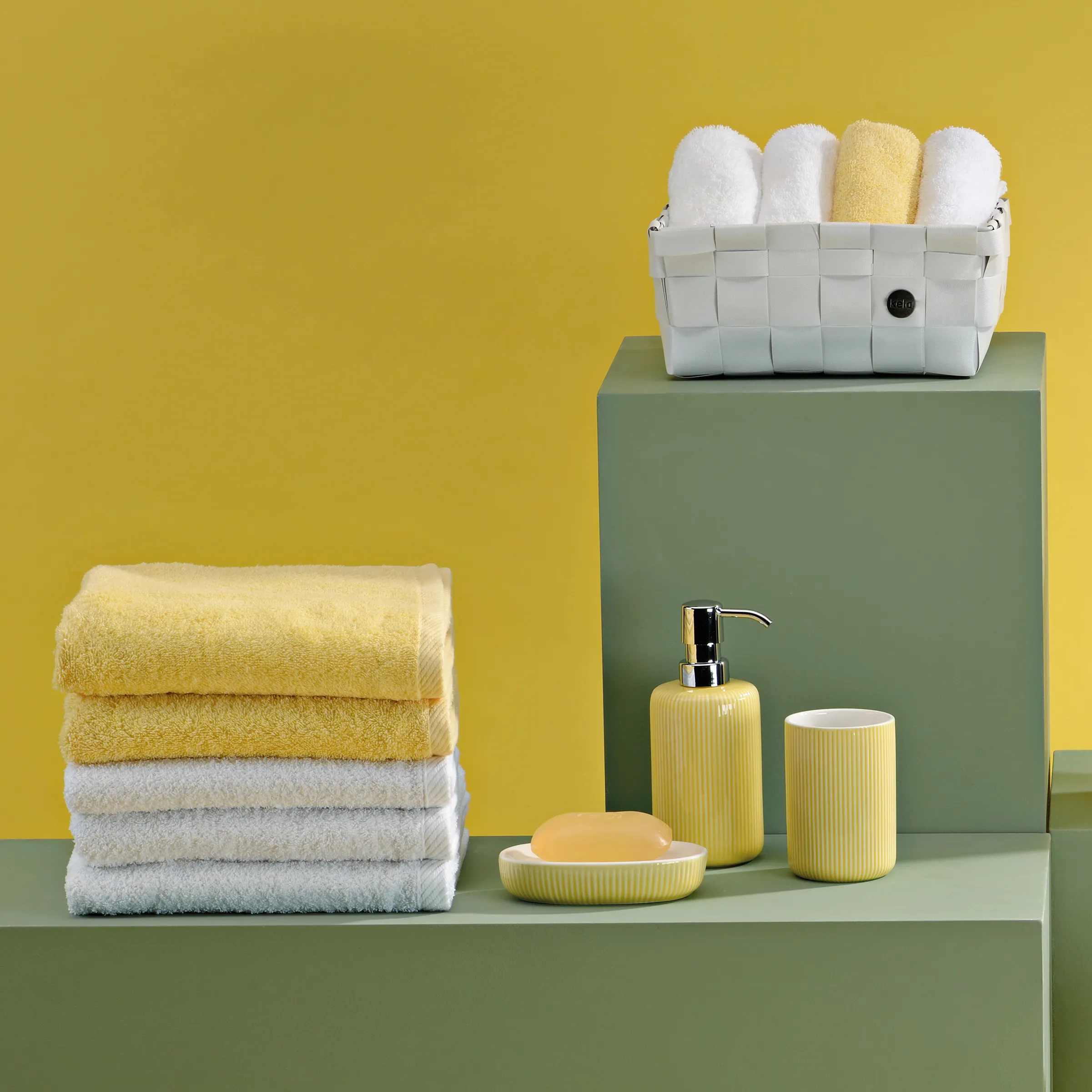 Badezimmer Accessoires Set - Handtücher und Seifenspender in Gelb Badezimmer-Set mit gelben Handtüchern, weißem Korb und gelbem Seifenspender auf grünem Podest