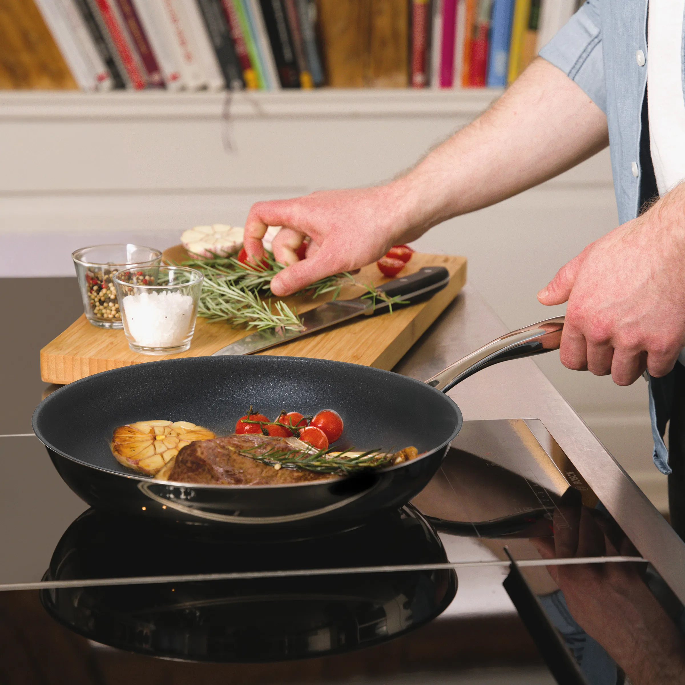 Steak braten in antihaftbeschichteter Pfanne – Kochen mit Rosmarin und frischen Kräutern Person brät Steak mit Rosmarin in beschichteter Pfanne, Schneidebrett mit Gewürzen und Kräutern im Hintergrund