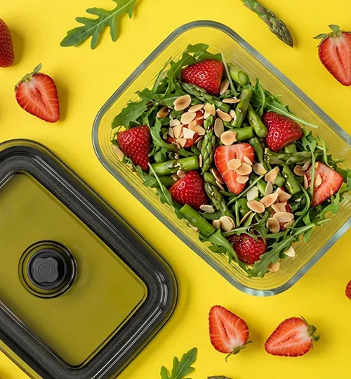 Gesunder Erdbeersalat mit Rucola und Mandeln in Meal Prep Dose Frischer Rucola-Salat mit Erdbeeren und Mandeln in einer Glas-Frischhaltedose auf gelbem Hintergrund