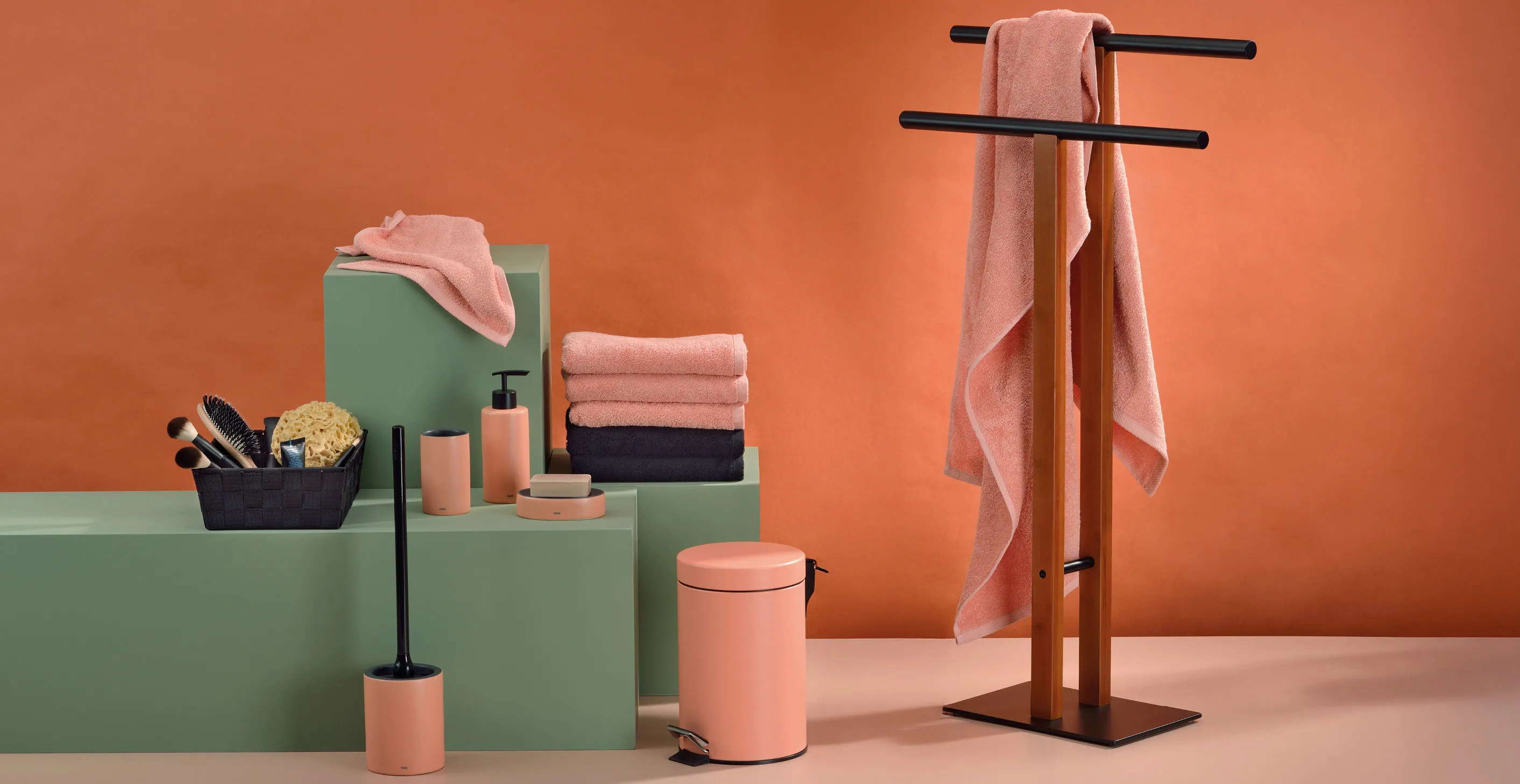 Badezimmer-Accessoires-Set mit rosa Handtüchern, Kosmetiktüchern, Seifenspendern und Holzhandtuchständer vor orange-grünem Hintergrund