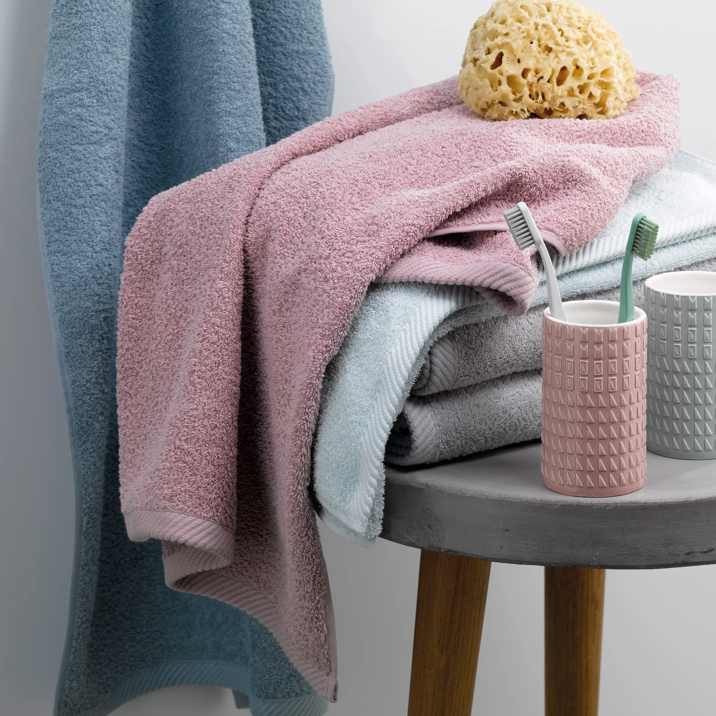 Badezimmer Handtücher und Accessoires Set Badezimmer-Arrangement mit gefalteten Handtüchern in Blau, Rosa und Weiß, Zahnbürsten in Behältern und gelbem Badeschwamm auf grauem Tisch