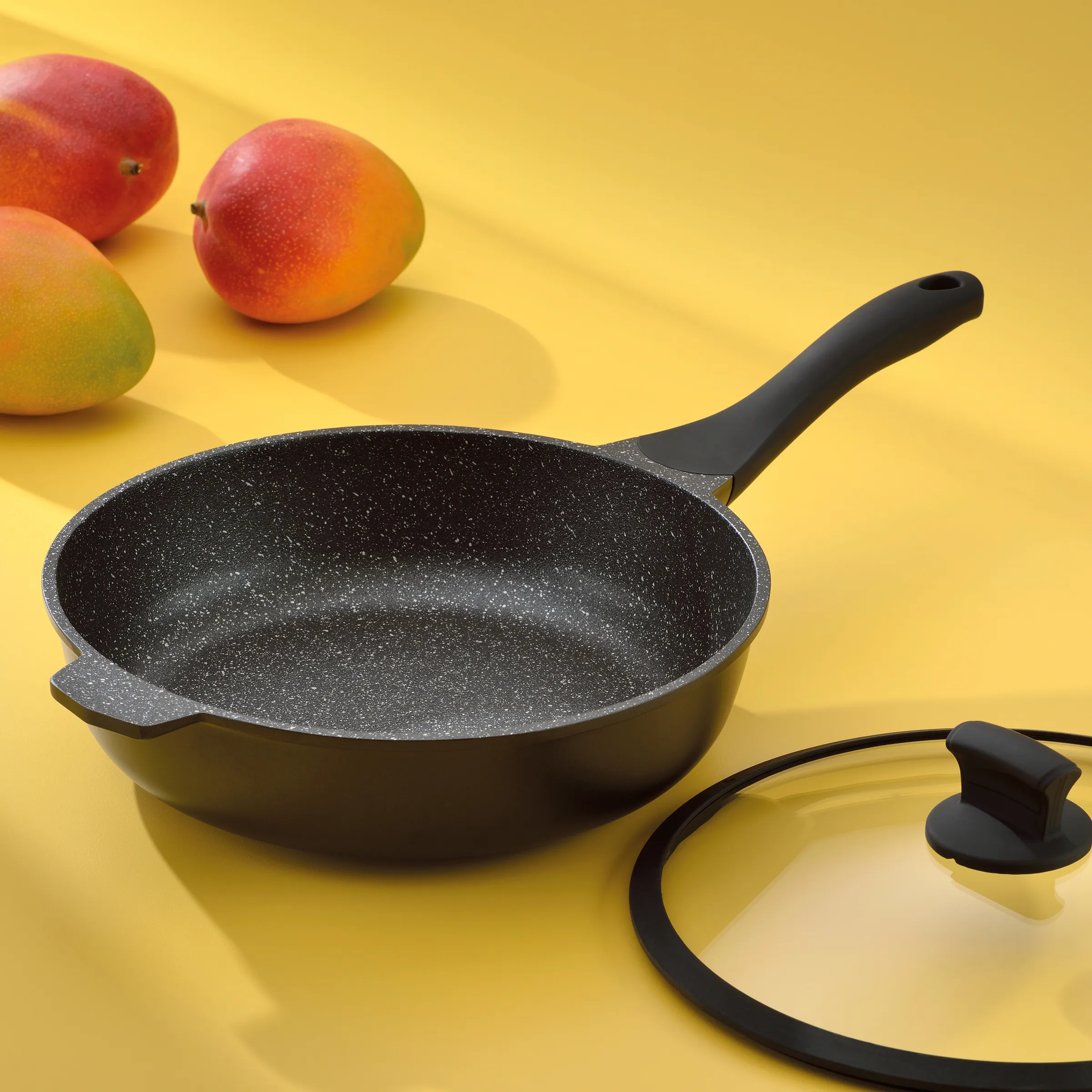Bratpfanne mit Antihaftbeschichtung und Glasdeckel - Hochwertige Kochpfanne Schwarze Bratpfanne mit Granitbeschichtung und Glasdeckel neben Mangos auf gelbem Hintergrund