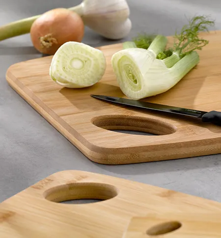 Schneidebrett aus Holz mit Löchern zur Aufbewahrung neben aufgeschnittener weißer Zwiebel und Dill mit Messer.