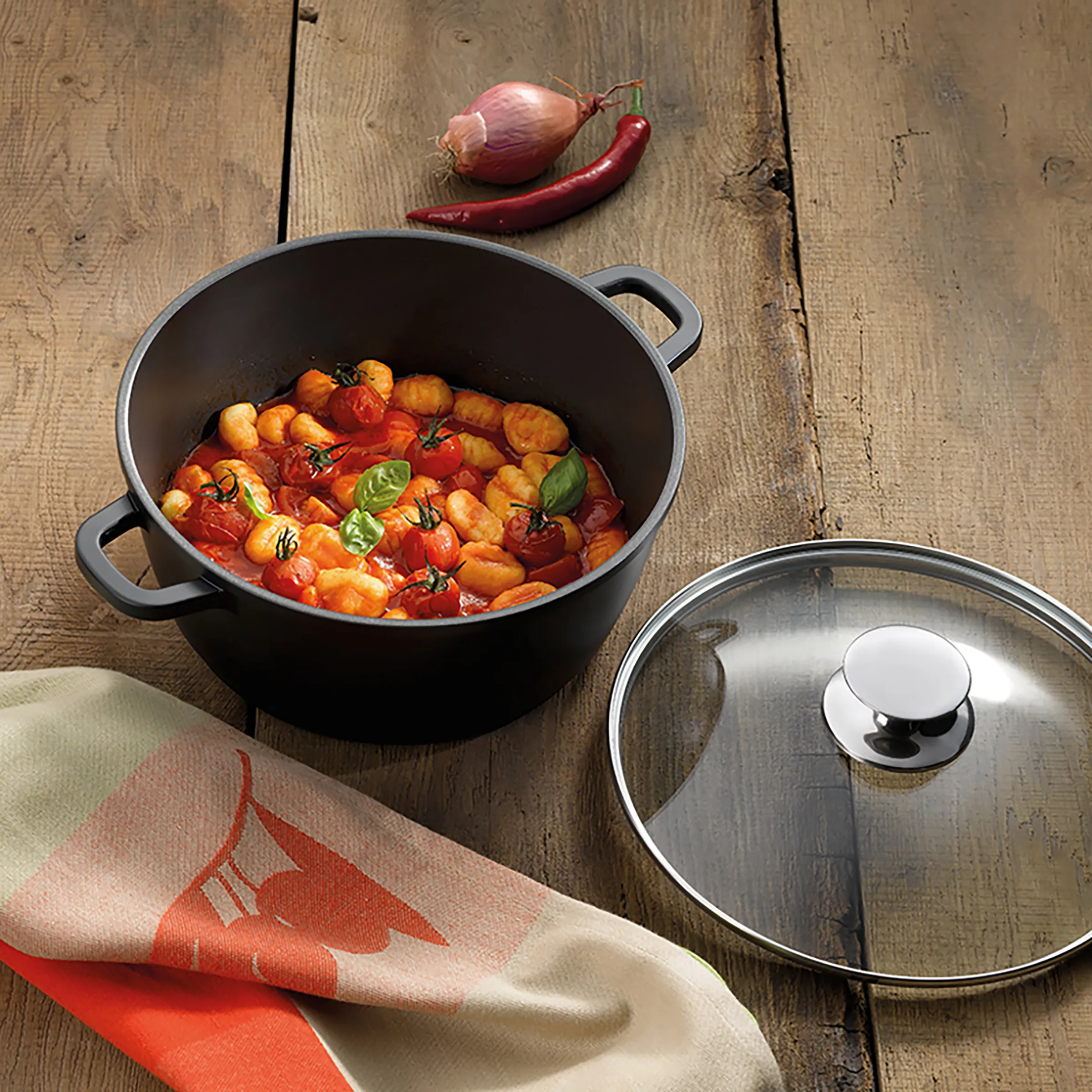 Kochtopf mit Deckel - Gnocchi in Tomatensauce zubereiten Schwarzer Kochtopf mit Gnocchi in Tomatensauce und Basilikum, Glasdeckel daneben, Schalotte und Chili auf Holztisch