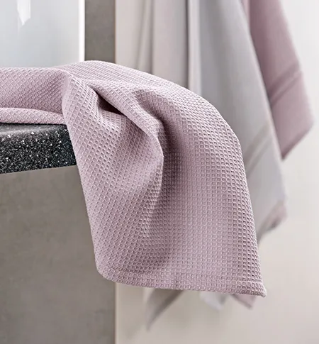 Hellrosa Waffelpiqué-Handtuch draped über grauem Behälter mit weiteren Handtüchern im Hintergrund