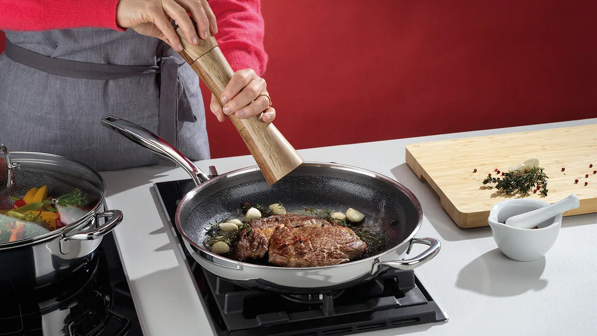 Person in rosafarbener Bluse würzt Fleisch in einer Pfanne auf dem Herd mit Pfeffer aus einer Mühle