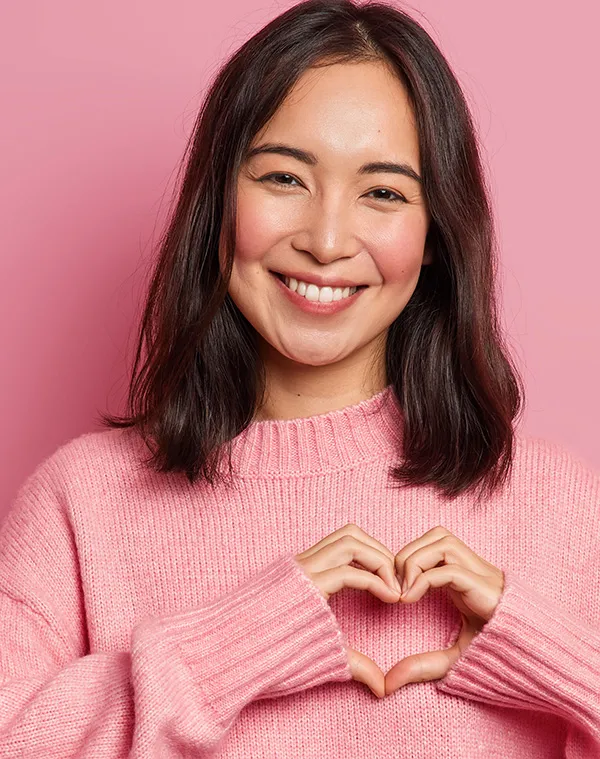 Junge Frau Herz-Geste rosa Pullover Portrait Lächelnde junge Frau mit langen dunklen Haaren in rosa Strickpullover zeigt Herz-Geste vor rosa Hintergrund
