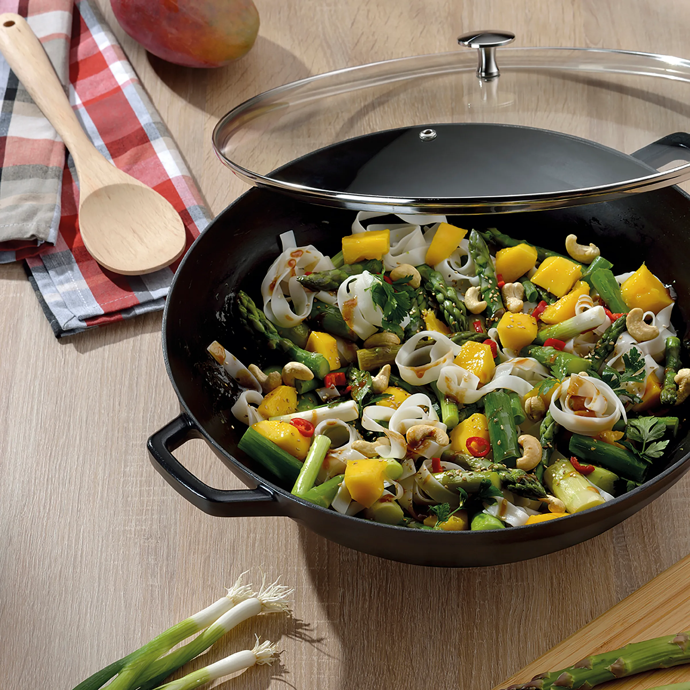 Wok-Pfanne mit Gemüse-Nudeln: Gesundes Pfannengemüse mit Spargel und Cashews Wok-Pfanne mit buntem Gemüse-Nudel-Gericht, Spargel, Paprika, Cashewnüssen und Glasdeckel auf Holztisch