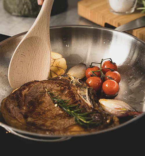Gebratenes Steak Rezept in der Pfanne mit Gemüse Gebratenes Fleischsteak in einer Pfanne mit Tomaten, Kartoffeln, Knoblauch und frischem Rosmarin, Holzlöffel sichtbar.