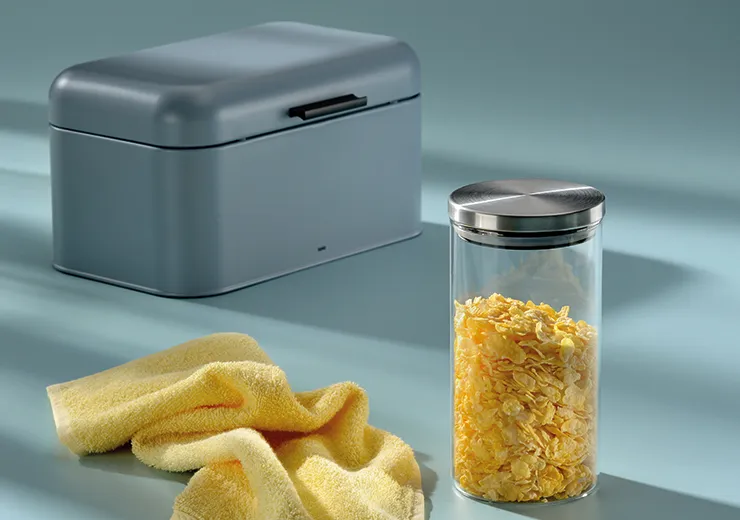 Graue Aufbewahrungsbox mit Deckel neben gelben Handtüchern und Glas mit Getreideflöckchen auf blauem Hintergrund
