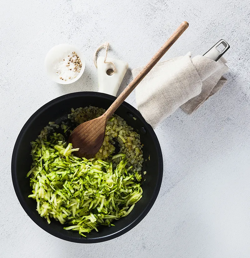 Zucchini raspeln und zubereiten - gesundes Gemüse kochen in der Pfanne Schwarze Pfanne mit geraspelten Zucchini und Holzlöffel, daneben Gewürze und Küchentuch auf heller Arbeitsfläche