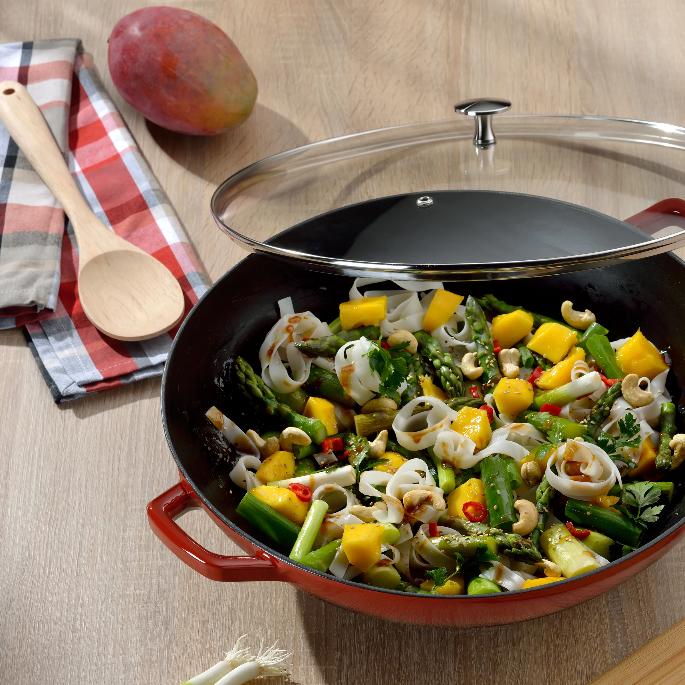 Wok-Pfanne mit Gemüse und Mango - Asiatische Küche Rote Pfanne mit Gemüse-Wok-Gericht aus Mango, Spargel, Zucchini und Cashewnüssen auf Holztisch