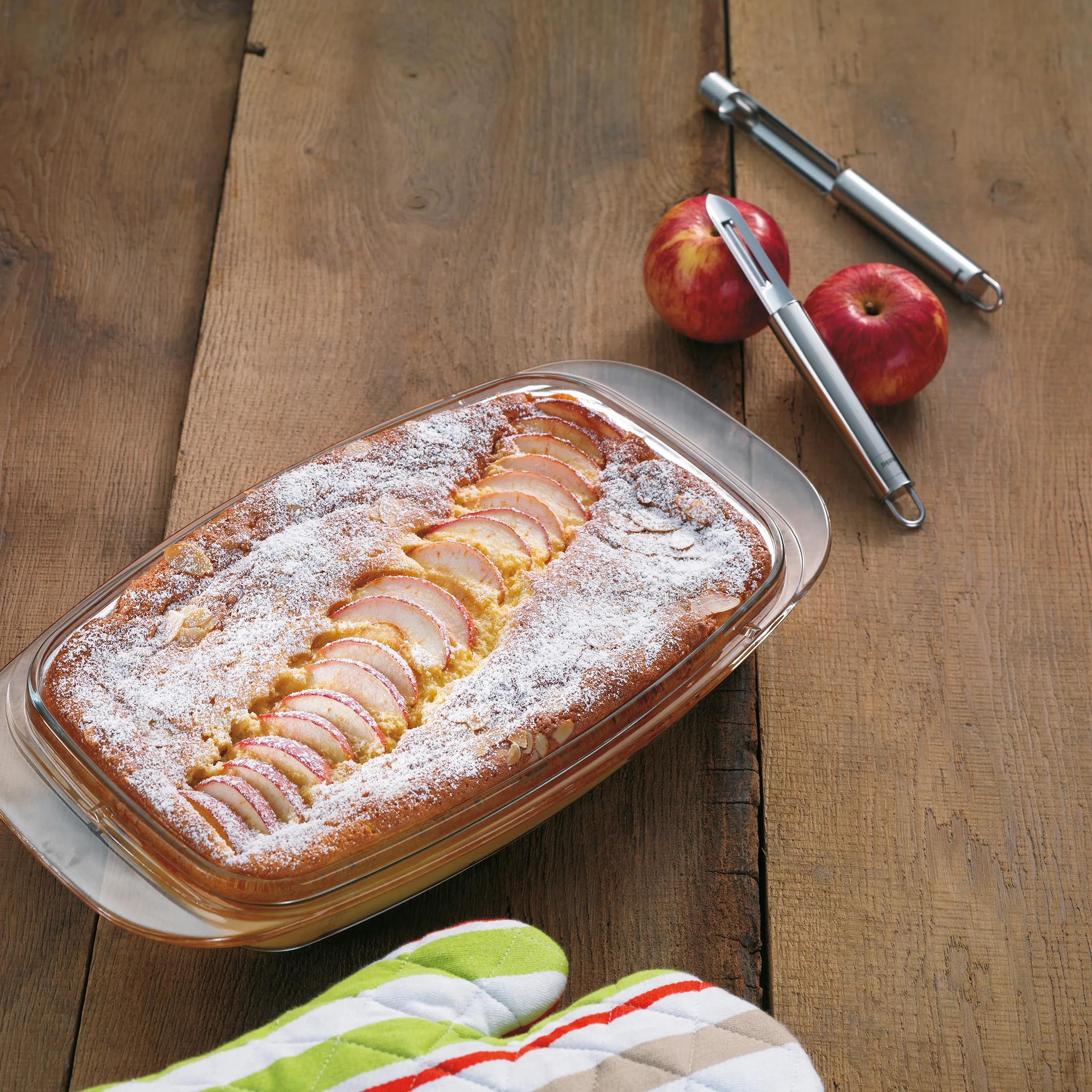 Apfelkuchen mit Puderzucker in Glasform auf Holztisch, daneben frische Äpfel und Küchenutensilien