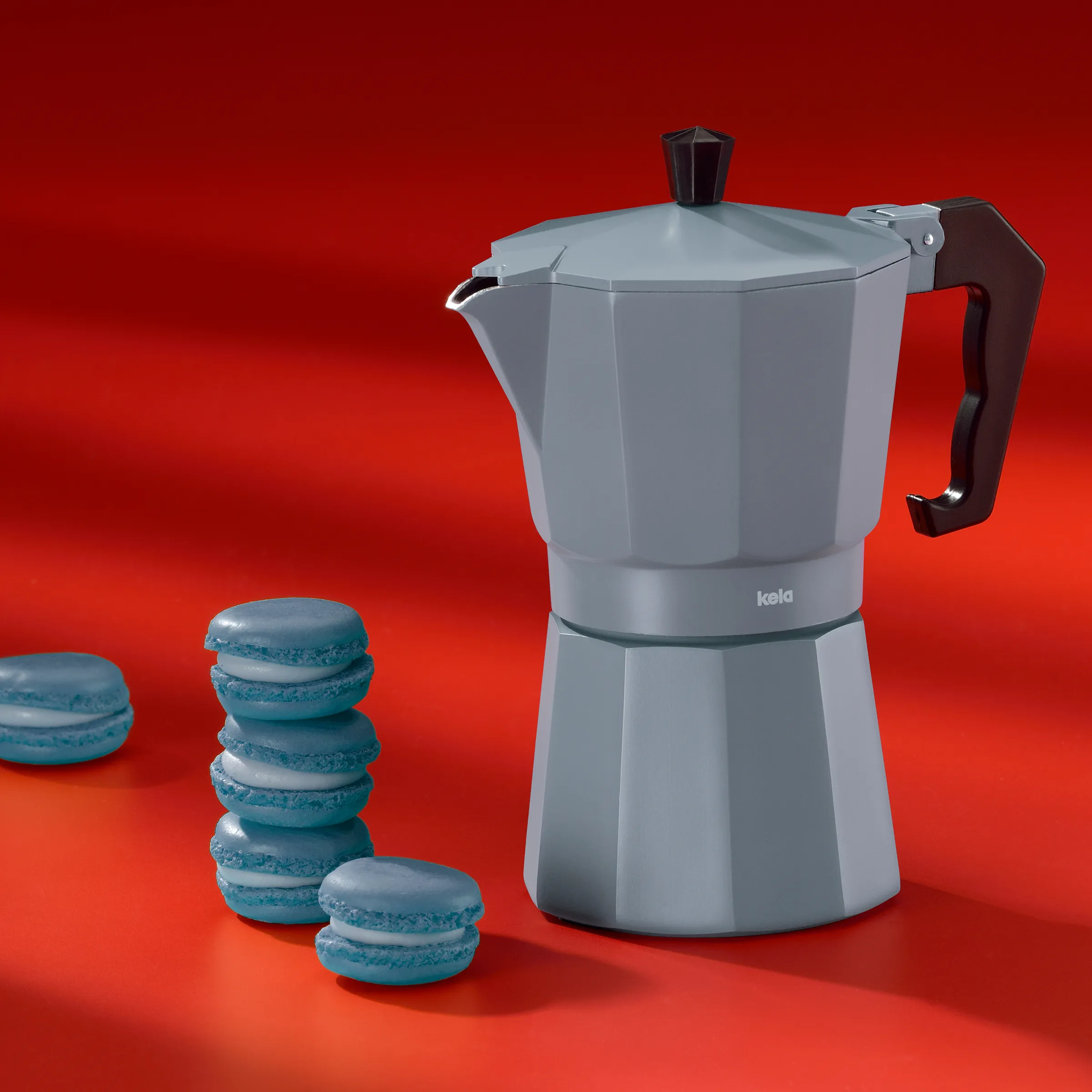 Keka Espressokocher Grau - Italienische Kaffeemaschine mit Macarons Keka Espressokocher in Grau mit schwarzem Griff neben gestapelten blauen Macarons vor rotem Hintergrund