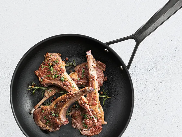 Lammkoteletts braten in Pfanne mit Rosmarin - Fleischzubereitung Gebratene Lammkoteletts mit Rosmarin in schwarzer Pfanne auf weißem Hintergrund
