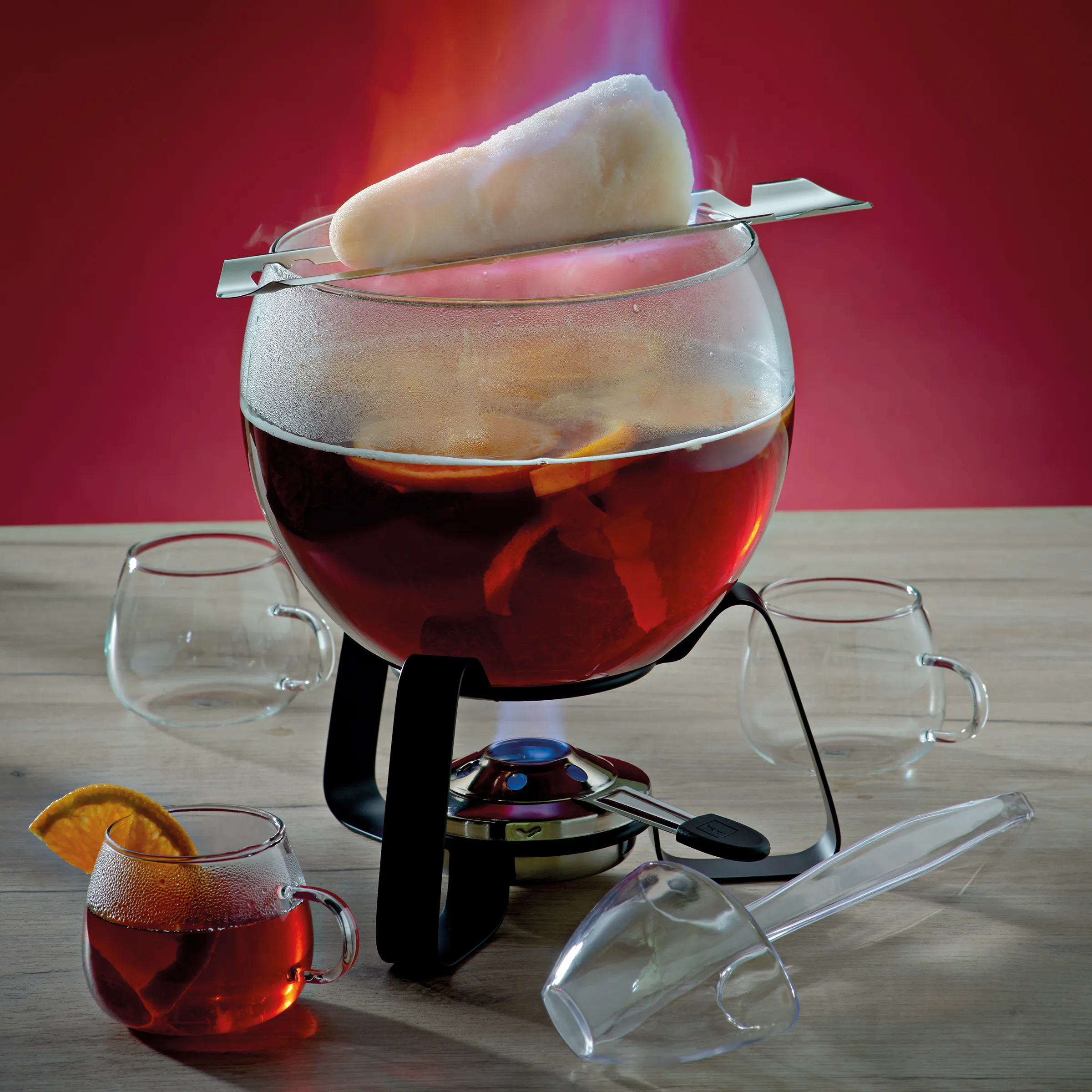Fondue Set - Fondue Pot mit Zubehör und Stövchen Fondue-Set mit großem Topf gefüllt mit roter Flüssigkeit, Stövchen, leeren Gläsern und Zangen auf Holztisch vor rotem Hintergrund