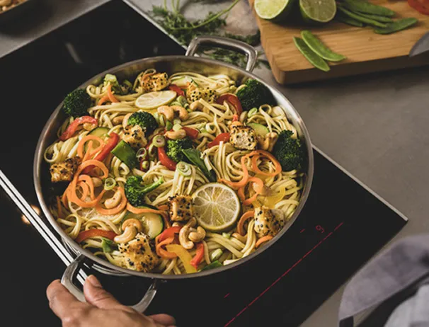 Asiatische Nudelpfanne mit Gemüse und Garnelen - gesundes Wok-Gericht Asiatische Nudelpfanne mit Gemüse, Garnelen und Sesam in Edelstahlpfanne auf Induktionsherd