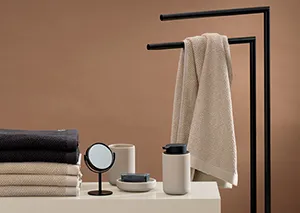 Modernes Badezimmer-Set mit schwarzem Handtuchständer, beigen Handtüchern und weißen Kosmetikprodukten auf neutralem Hintergrund