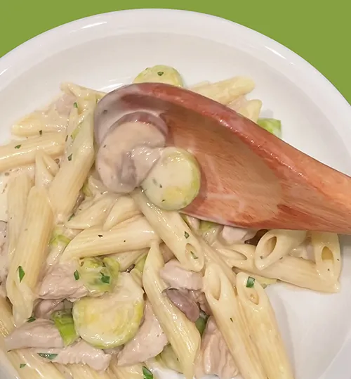 Pasta-Gericht mit Penne, Zucchini und Fleisch in weißer Schale mit Holzlöffel auf grünem Hintergrund