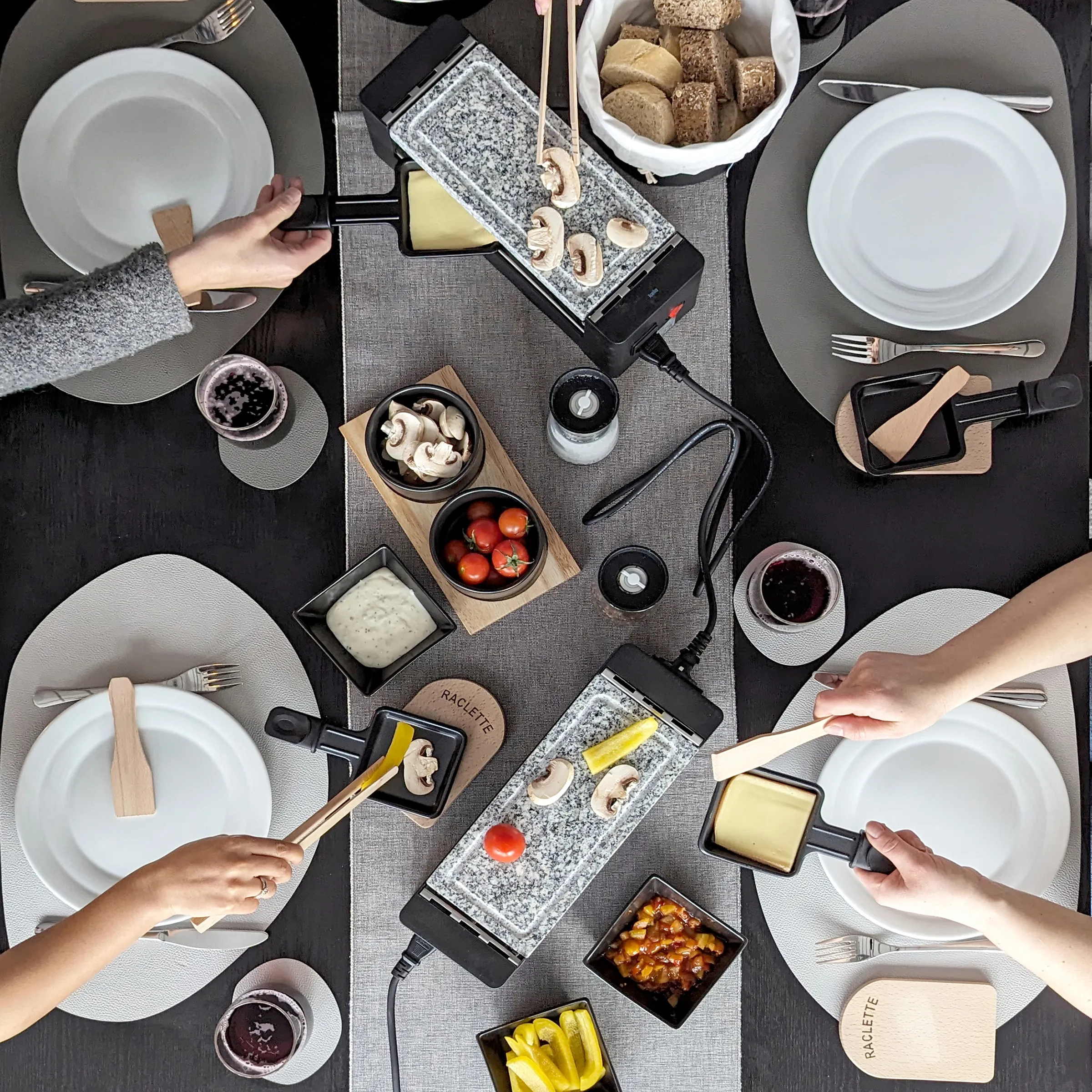Raclette-Dinner mit vier Personen an rundem Tisch, Käse auf Grillplatten, frische Pilze, Tomaten und Brot