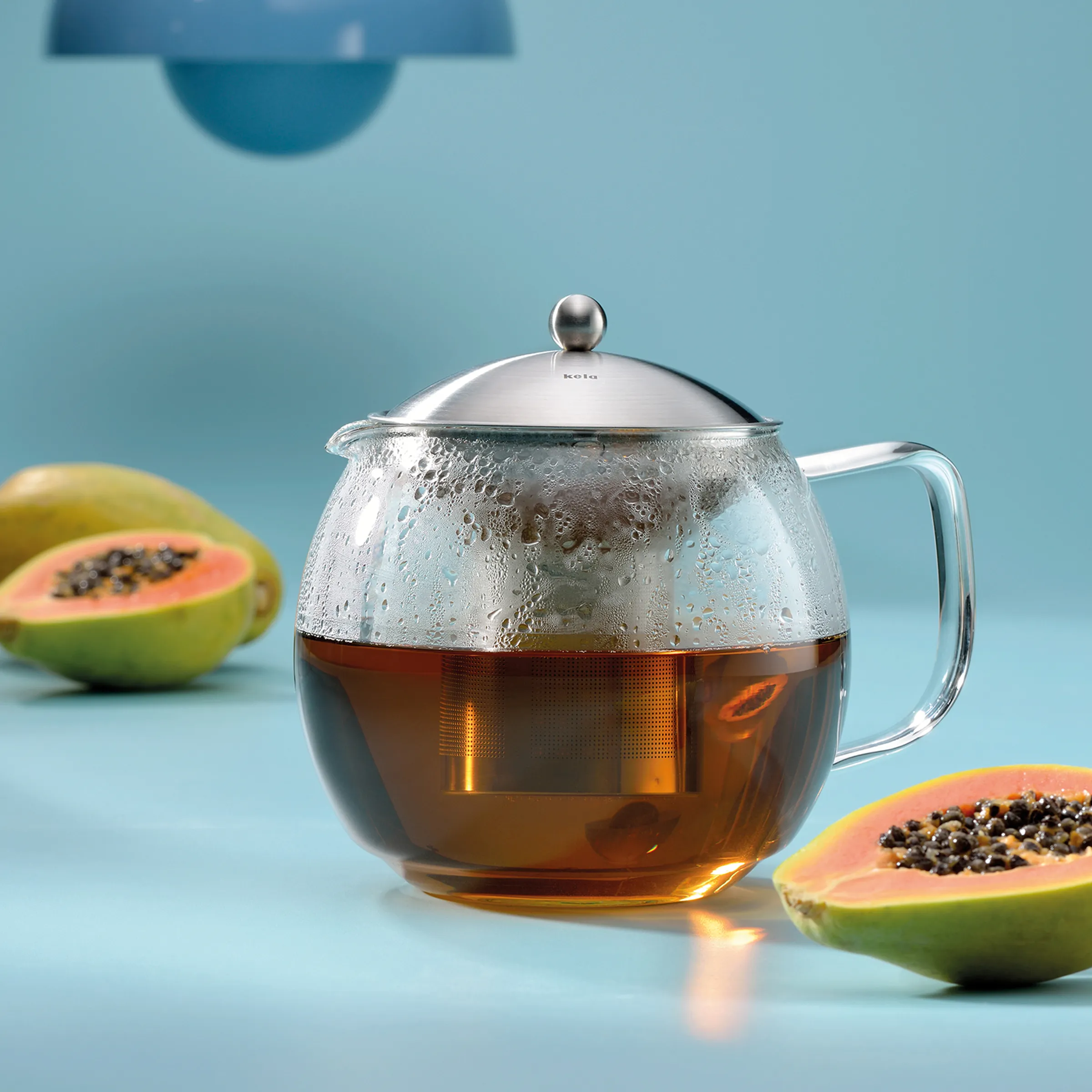 Glas-Teekanne mit Edelstahldeckel gefüllt mit goldbraunem Tee, daneben aufgeschnittene Papaya auf blauem Hintergrund