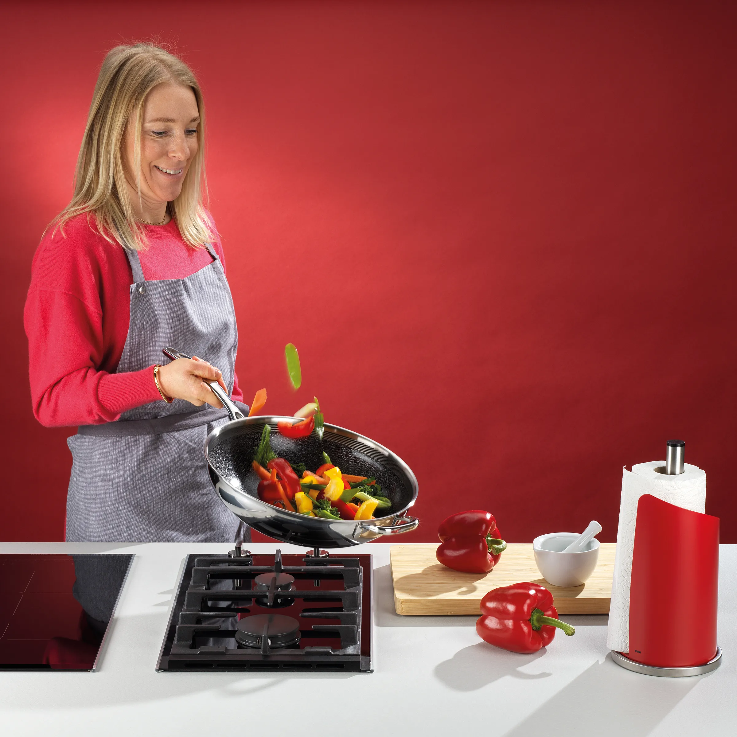 Wok Kochen mit frischem Gemüse - Paprika braten Frau in roter Jacke und grauer Schürze brät bunte Paprika in einem Wok auf einem Gasherd vor rotem Hintergrund