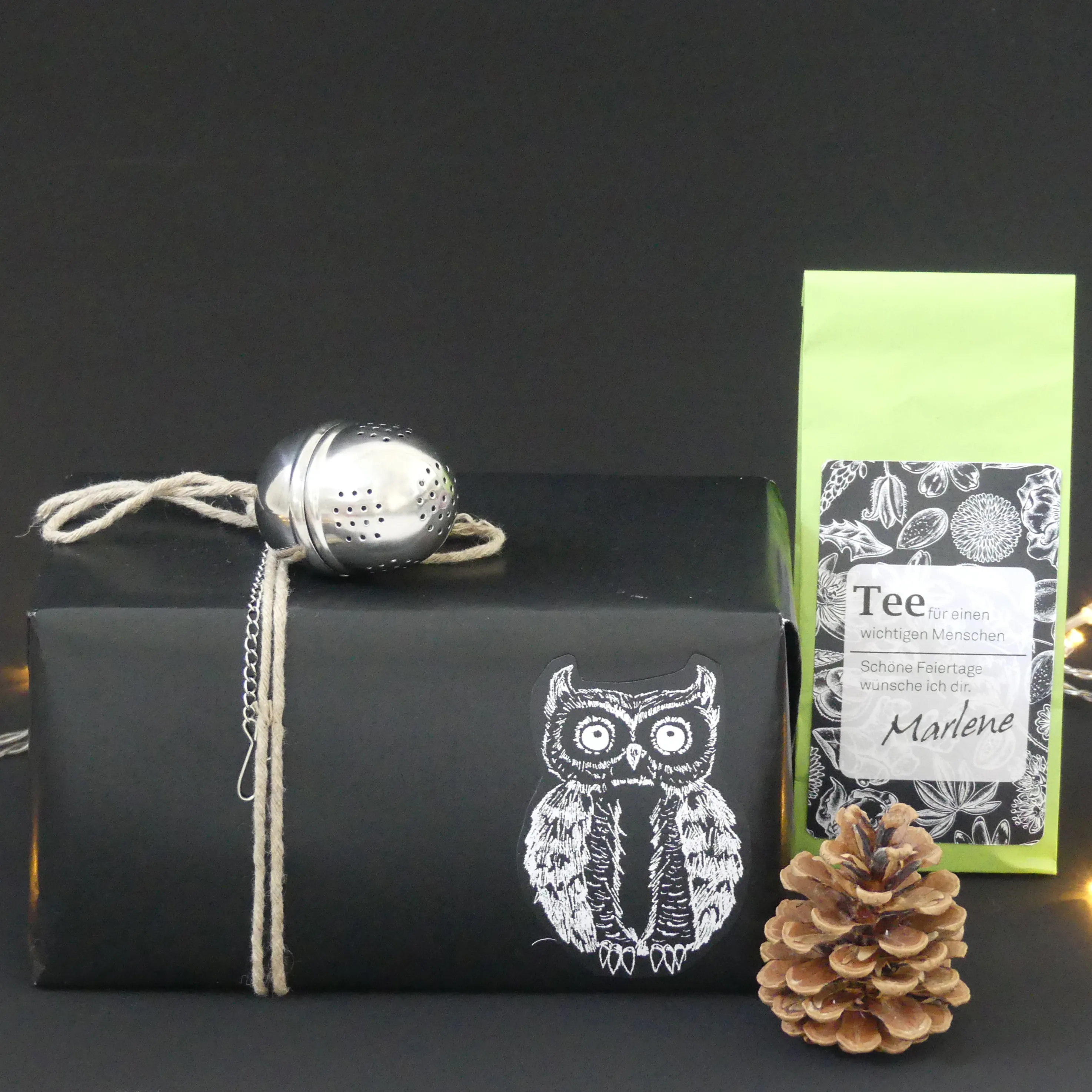 Schwarze Geschenkbox mit Eulen-Motiv, Tee-Verpackung mit grünem Label und silbernem Teeei auf Schnur neben Tannenzapfen