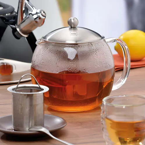 Teekanne Glas mit Sieb - Tee Zubereitung Glaskanne mit heißem Tee und Metallsieb auf Holztisch mit Zitrone und Tasse im Hintergrund