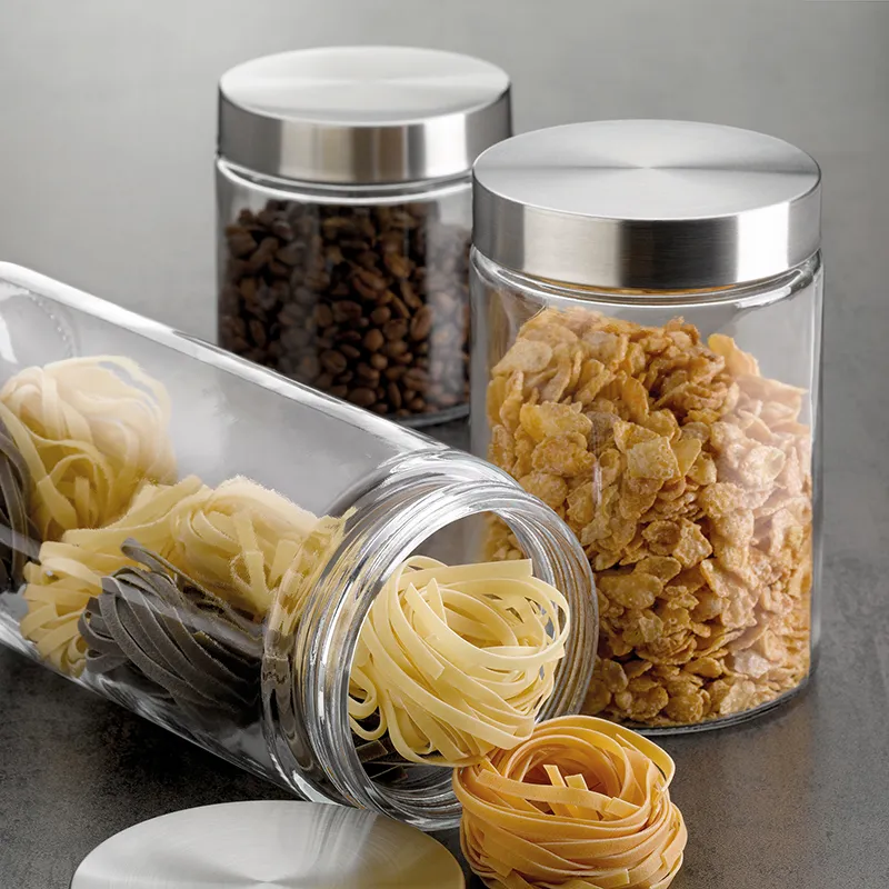 Drei Vorratsgläser mit Metalldeckel gefüllt mit Nudeln, Cornflakes und Kaffeebohnen auf dunkler Arbeitsfläche