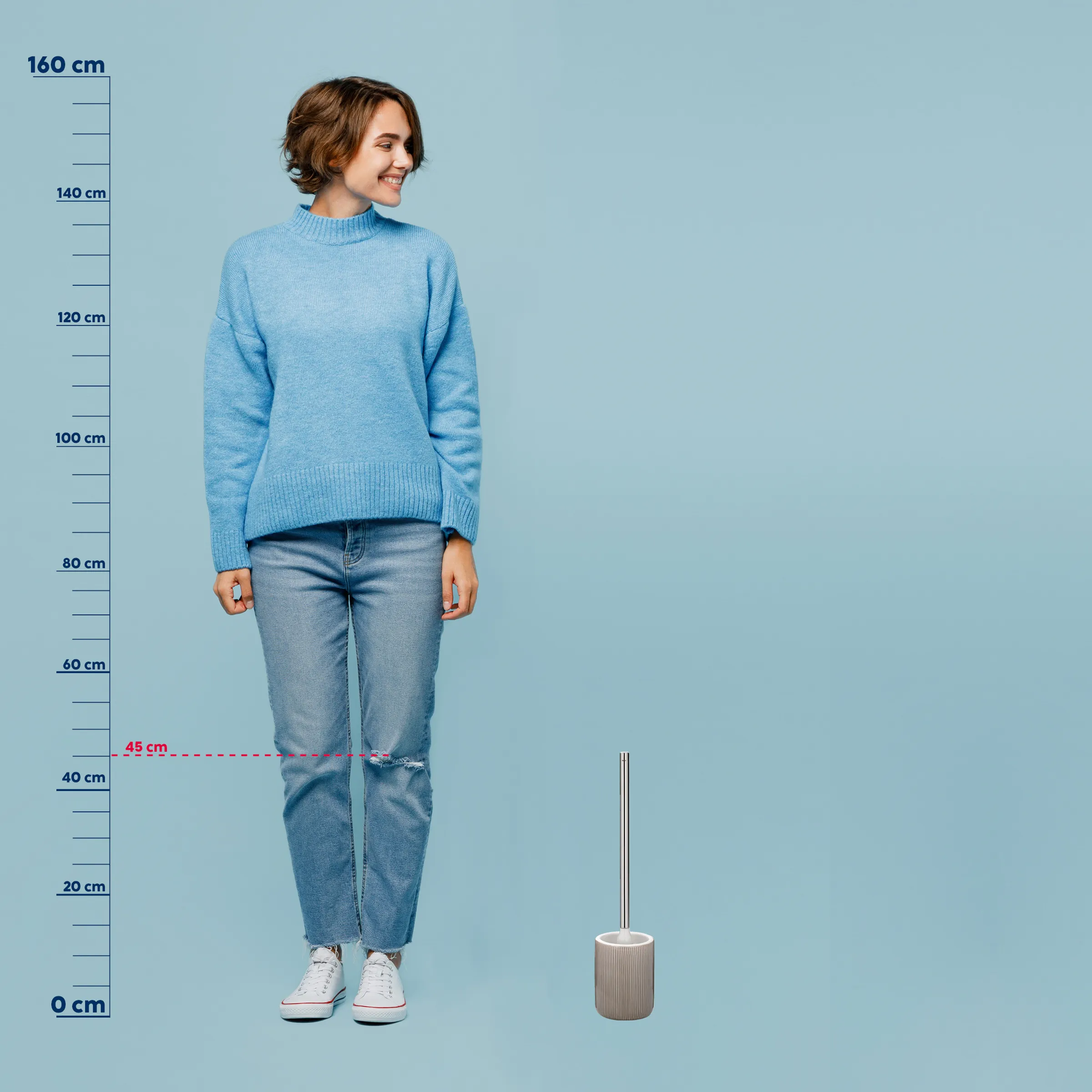 Größenvergleich: Badezimmerbecher mit Höhenskala und Frau Junge Frau in hellblauem Strickpullover und zerrissener Jeans steht vor Größenskala, neben Badezimmerbecher.