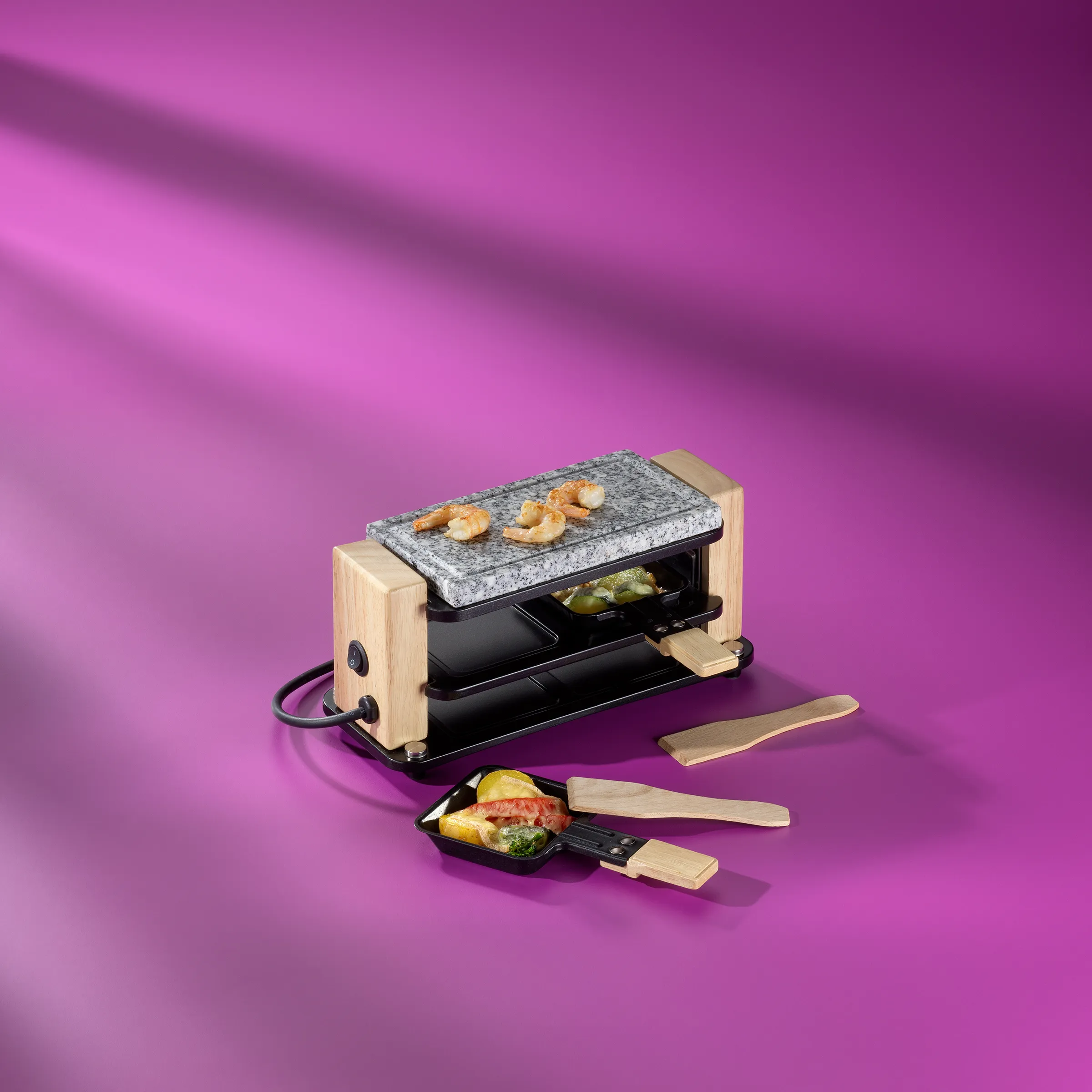 Raclette-Grill mit Holzgehäuse und Granitstein, zwei Pfännchen mit Gemüse und Käse auf violettem Hintergrund