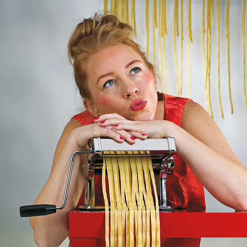 Frau macht frische Pasta mit Nudelmaschine - Hausgemachte Nudeln Blonde Frau in rotem Kleid mit Nudelmaschine, Pasta wird hergestellt, gelbe Fettuccine hängen im Hintergrund