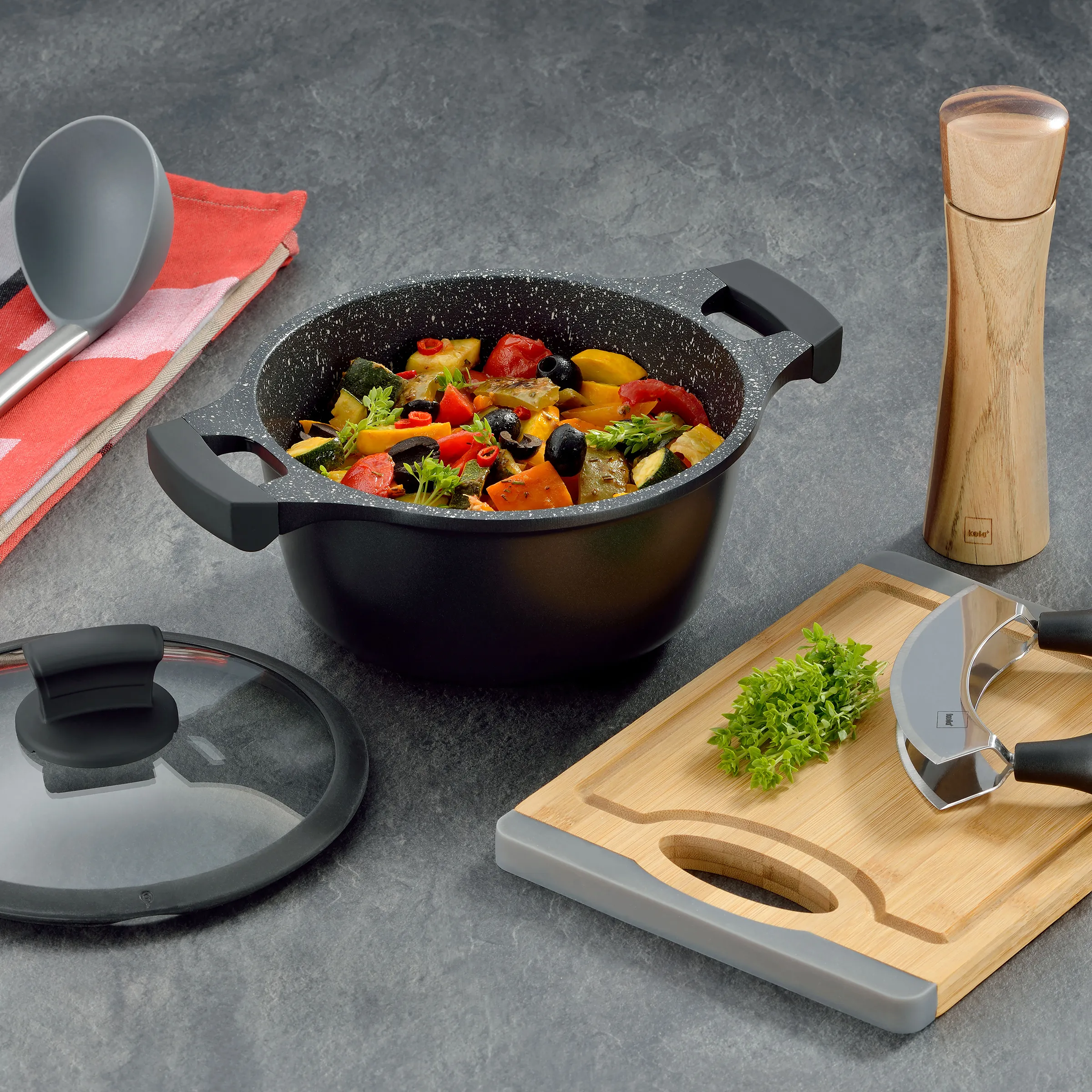 Kochset mit Topf, Schneidebrett und Küchengeräten Schwarzer Kochtopf mit Gemüse, Holzschneidebrett, Pfeffermühle und Küchenutensilien auf grauem Untergrund.