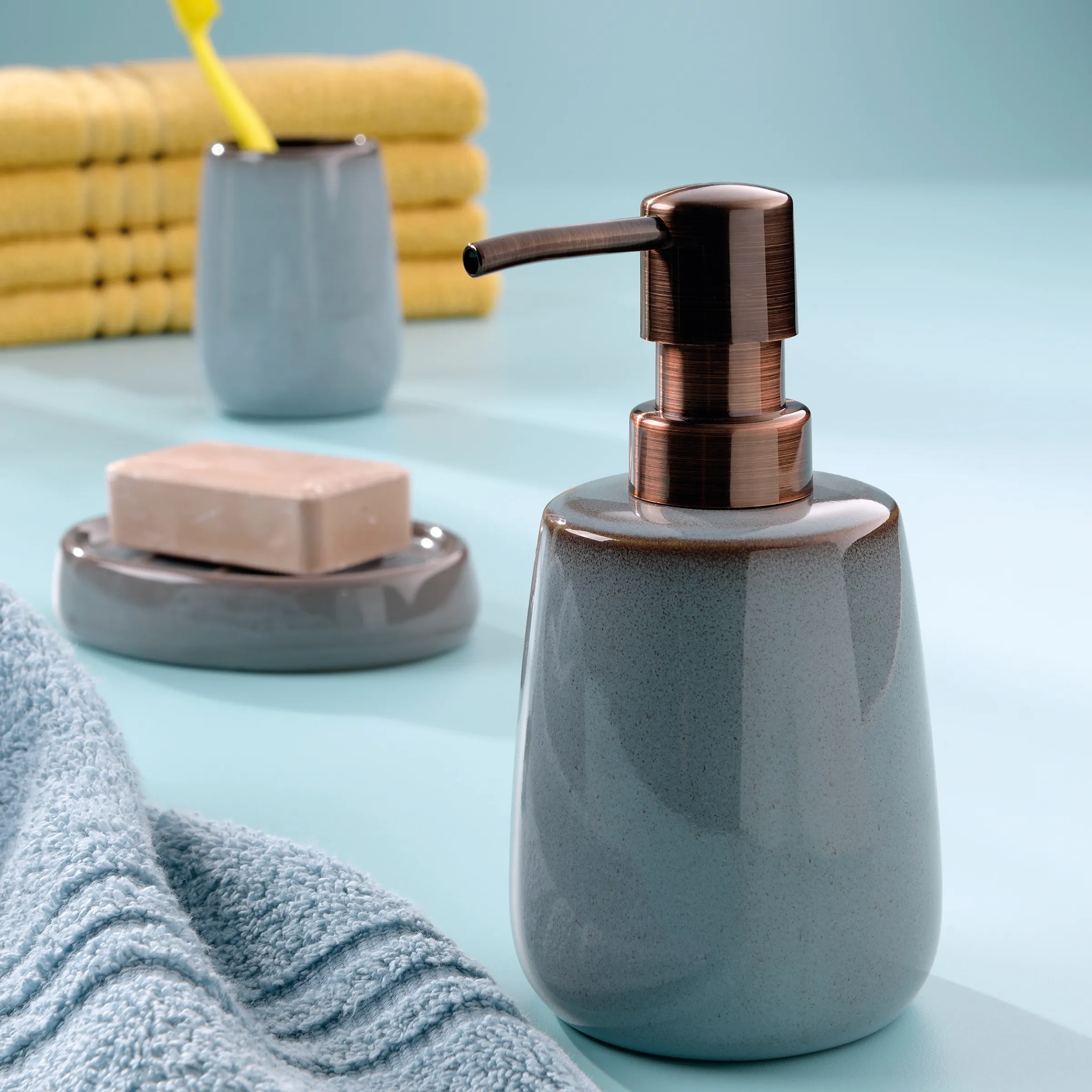 Badezimmer-Set mit grauem Seifenspender, Seifenschale, Handtüchern und gelben Badetüchern auf hellblauem Hintergrund.