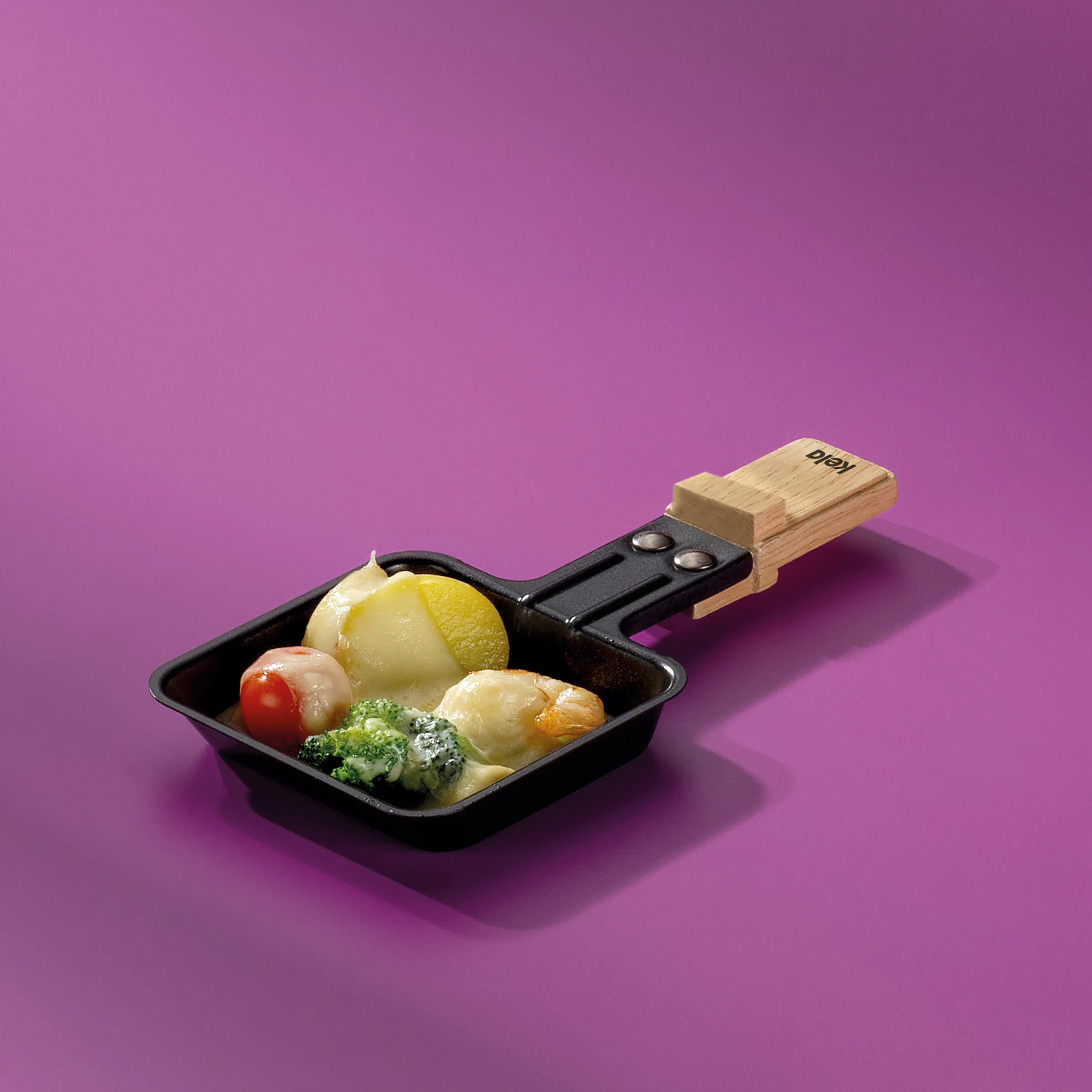 Raclette Pfanne - Mini Käse Pfannchen mit Gemüse Kleine schwarze Raclette-Pfanne mit Holzgriff gefüllt mit Kartoffel, Broccoli, Tomate und Käse auf violettem Hintergrund