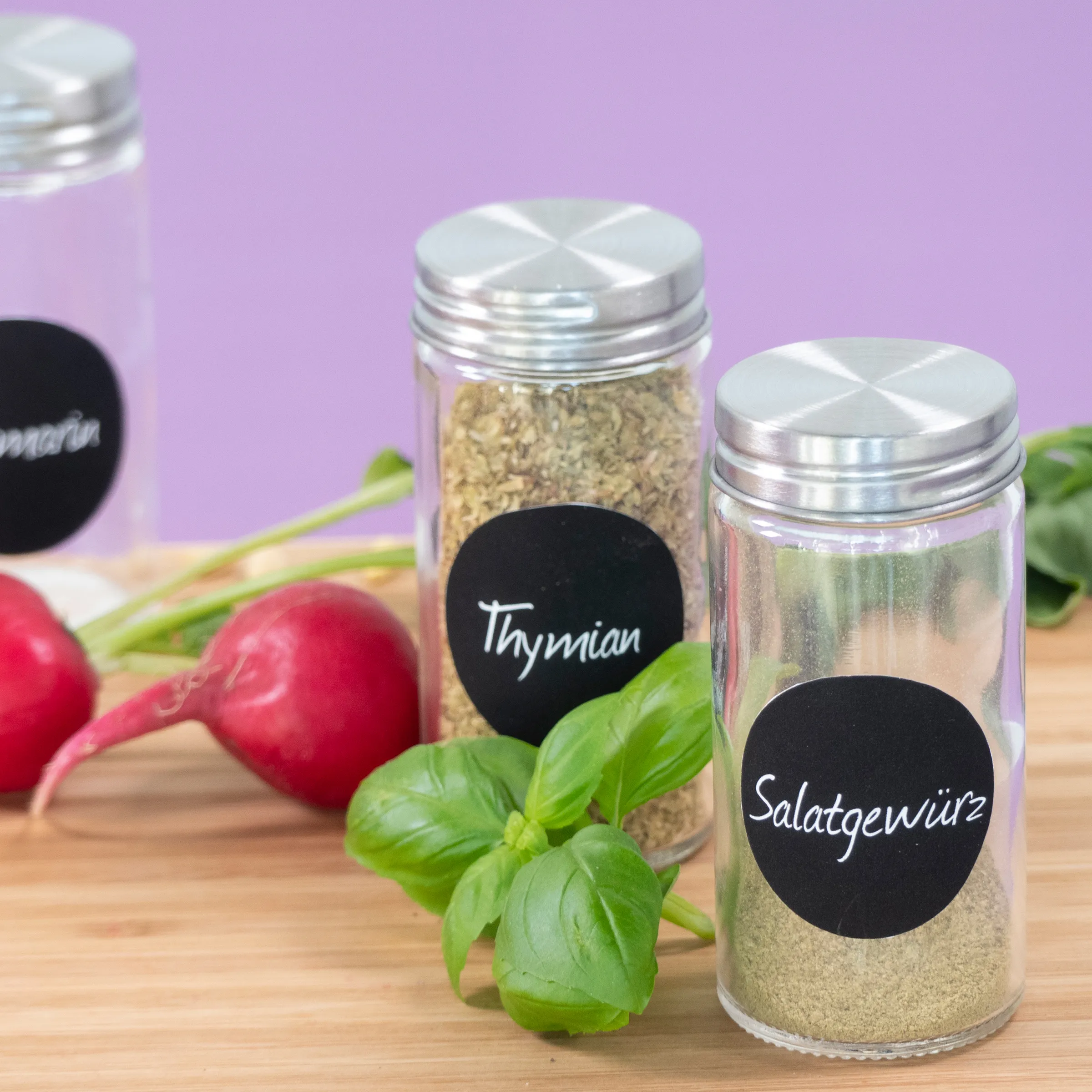 Gewürzgläser Thymian und Salatgewürz - Küchenlagerung Gewürzgläser mit Metalldeckeln auf Holzbrett mit frischen Kräutern und Radieschen vor lila Hintergrund