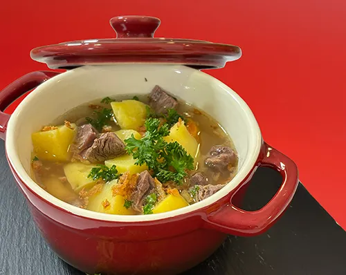 Rindfleischeintopf mit Kartoffeln im roten Emailletopf - Schmorgericht Roter Emailletopf mit Rindfleischeintopf, Kartoffeln und frischer Petersilie auf schwarzem Untergrund