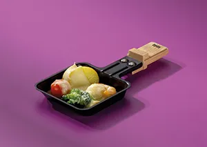 Raclette-Pfanne mit Käse und Kartoffeln - Schweizer Käsespezialität Schwarze Raclette-Pfanne mit geschmolzenem Käse, Kartoffeln und Tomate auf lila Hintergrund
