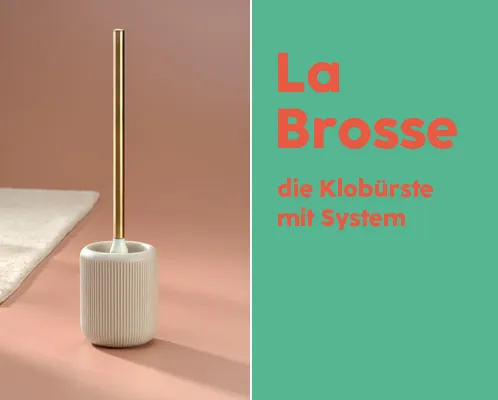 La Brosse Toilettenbürste mit System - Design WC-Bürste Weiße Toilettenbürste mit goldenem Stiel auf rosafarbenem Untergrund neben grünem Bereich mit deutscher Produktbeschreibung