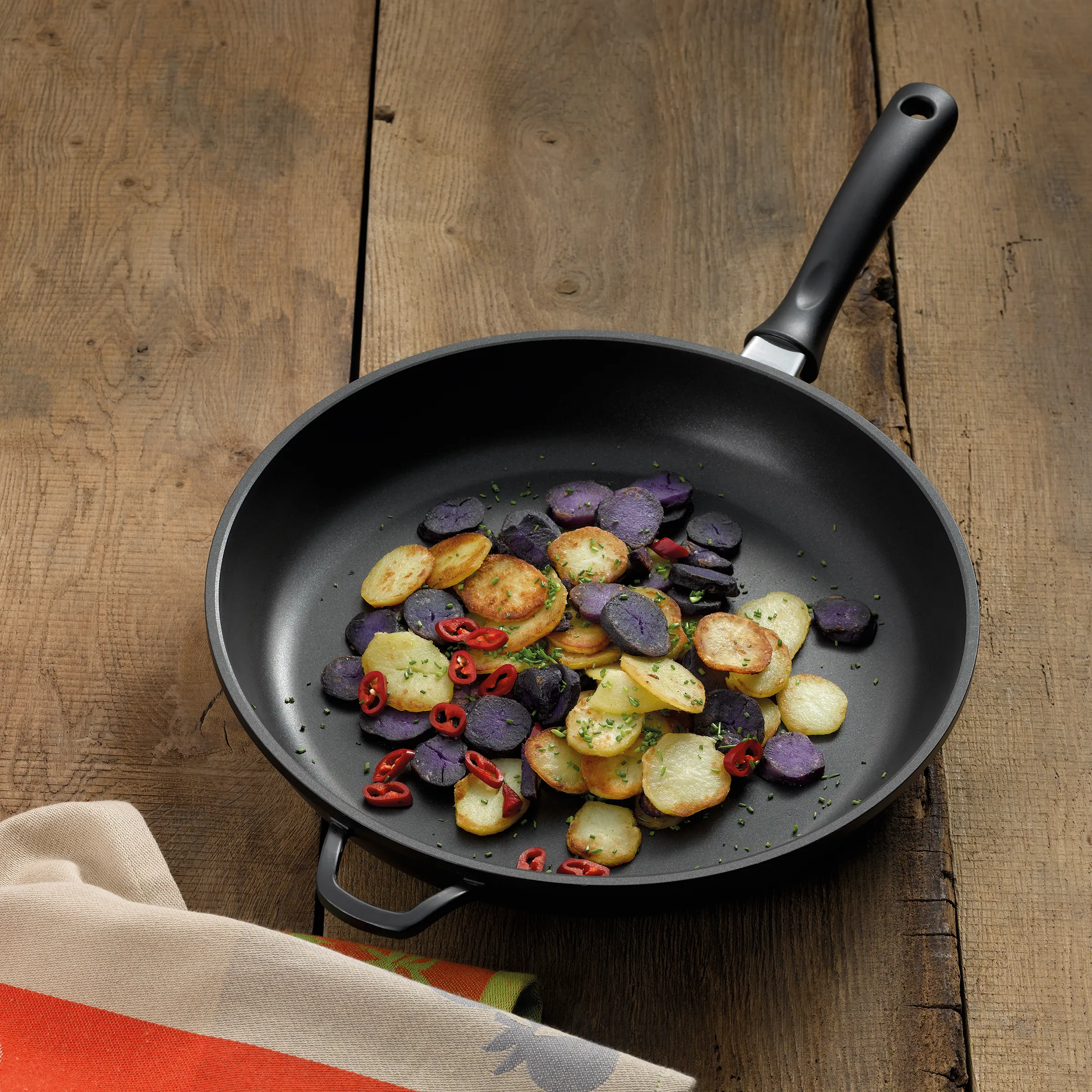 Bratpfanne mit Bratkartoffeln und buntem Gemüse - Antihaft-Pfanne Küchenzubehör Schwarze Bratpfanne mit gebratenen Kartoffelscheiben, lila Kartoffeln, roter Paprika und Kräutern auf Holztisch