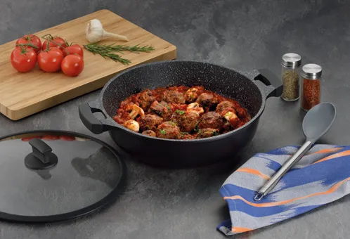 Fleischbällchen Topf - Eintopf Kochset mit Zubehör Schwarzer Topf mit Fleischbällchen-Ragout auf grauer Arbeitsplatte, daneben Schneidebrett mit Tomaten und Rosmarin