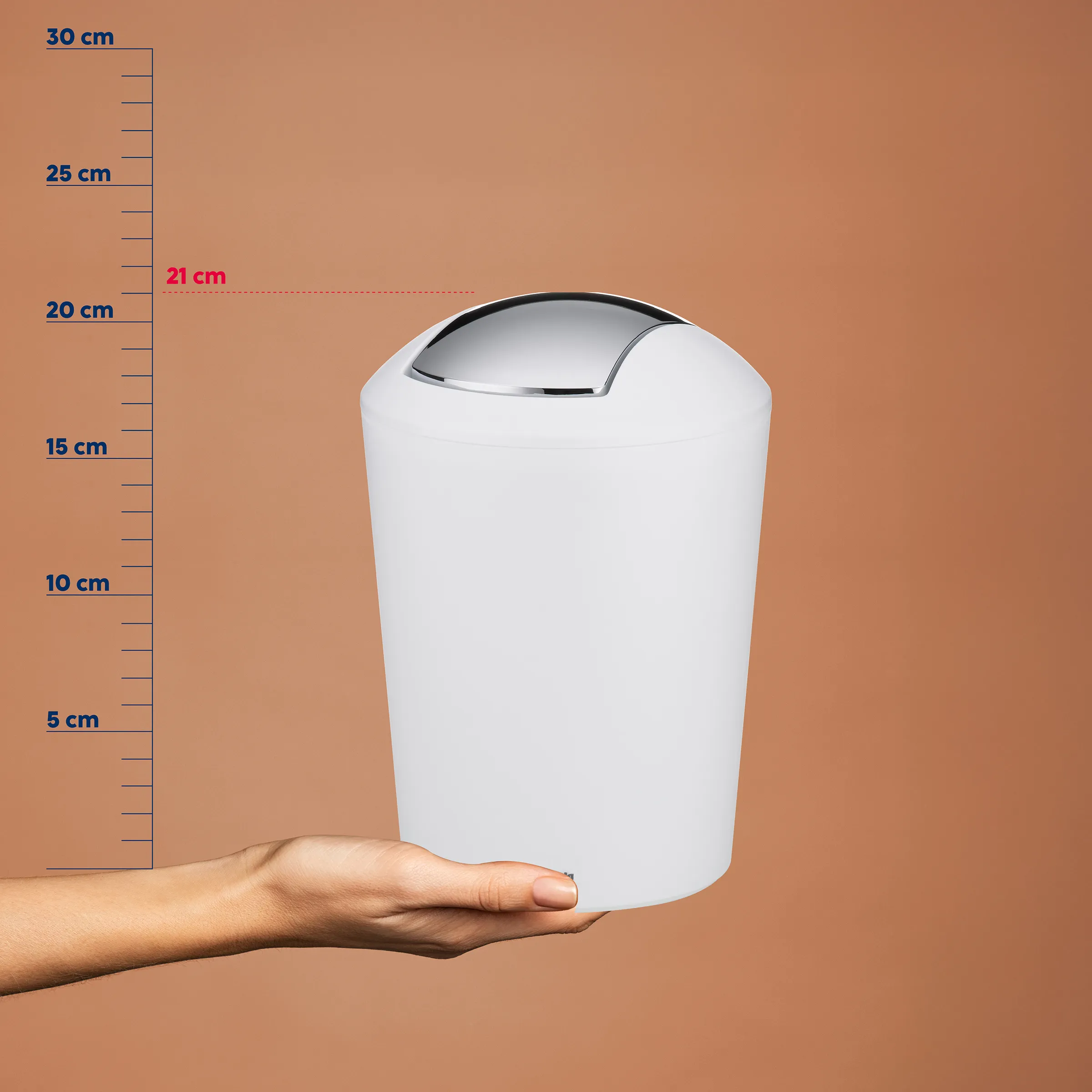 Weißer Schwingdeckel-Abfalleimer 21 cm - Kosmetikeimer Mülleimer Bad Weißer Schwingdeckel-Abfalleimer 21 cm hoch mit Größenskala, von Hand gehalten vor beigem Hintergrund