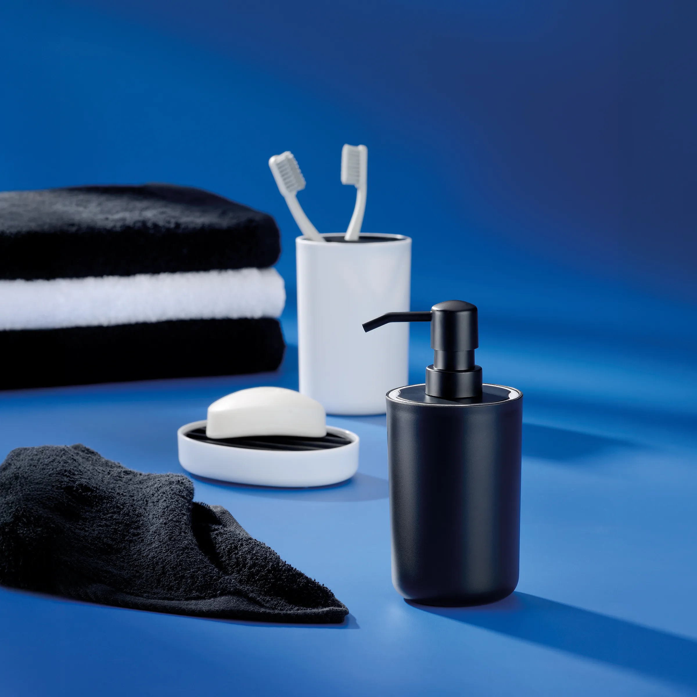 Schwarzes Seifenspender, weiße Seifenschale, Zahnputzbecher mit Zahnbürsten und schwarz-weiße Handtücher auf blauem Hintergrund
