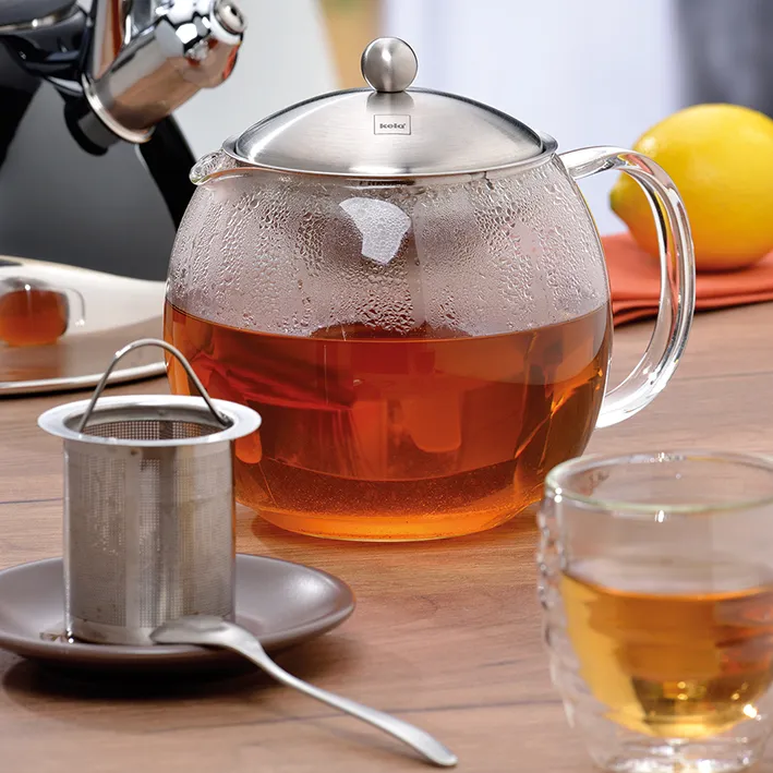 Glas-Teekanne mit Sieb und Edelstahl-Deckel für losen Tee Glas-Teekanne mit Edelstahl-Deckel gefüllt mit frisch gebrühtem Tee, daneben Tee-Ei und Teetasse auf Holztisch