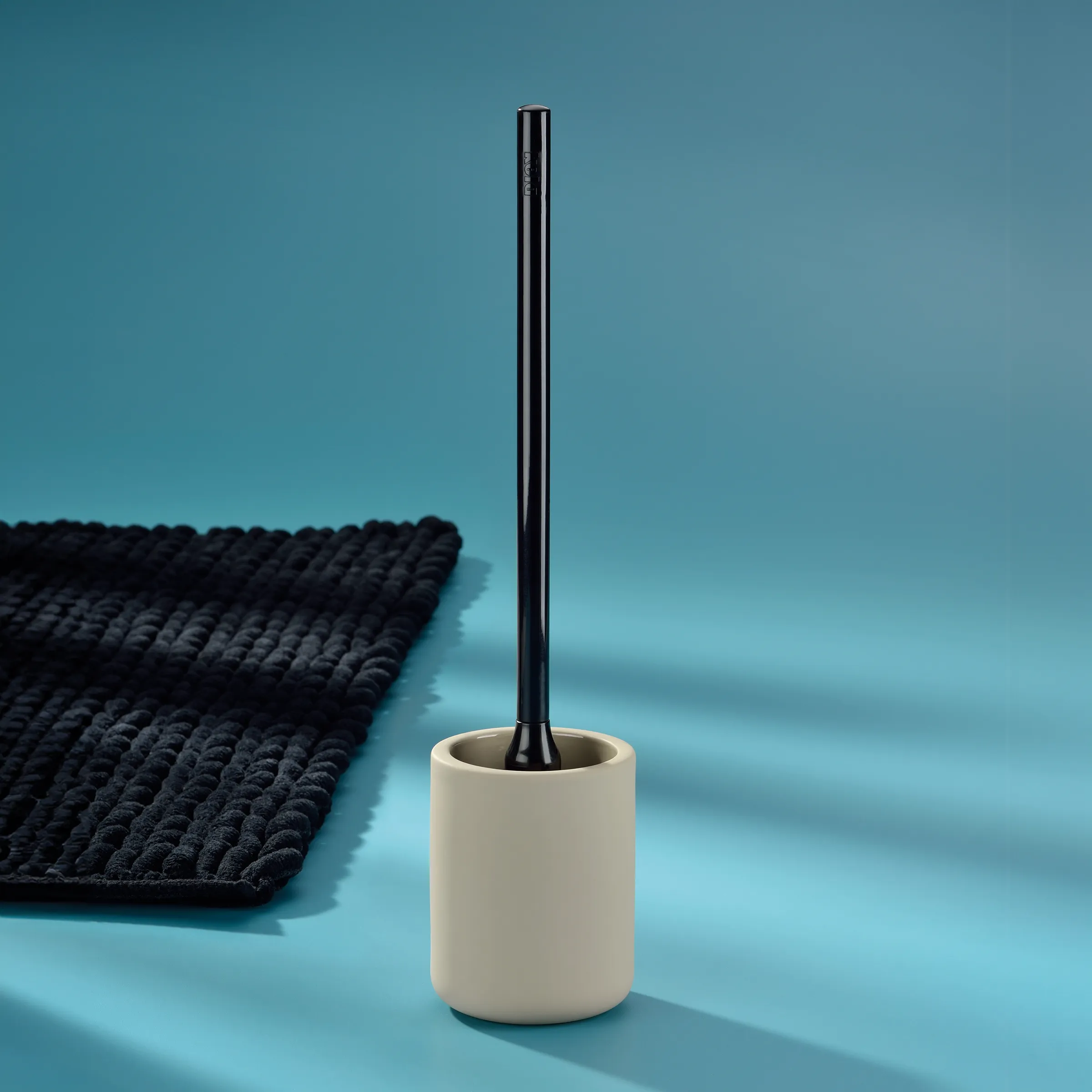 Toilettenbürste weiß mit Halter - Badaccessoire Cremeweiße Toilettenbürste mit schwarzem Griff und Behälter neben dunklem Badteppich auf blauem Hintergrund