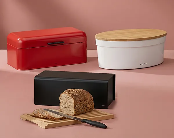 Drei moderne Brotboxen in Rot, Weiß und Schwarz mit Brotlaib und Schneidebrett vor rosa Hintergrund