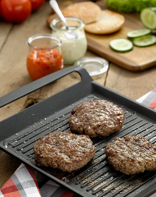 Hamburger Patties grillen - perfekte Grillrezepte Drei Hamburger-Patties braten auf schwarzem Grillgitter neben Tomaten, Gurken, Brötchen und Getränken