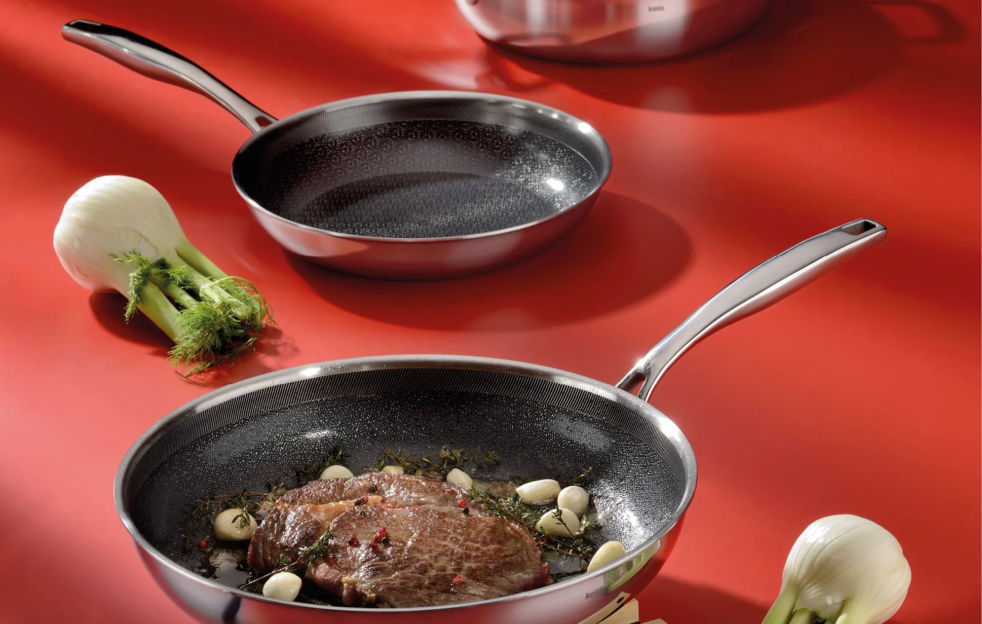 Zwei Edelstahl-Pfannen auf rotem Hintergrund: Eine mit gebratenem Steak, Knoblauch und Kräutern, eine leer. Daneben frisches Gemüse.